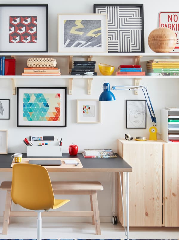 A desk on castors stands by a wall, surrounded by light-wood IVAR cabinets, wall shelves, and framed artwork.