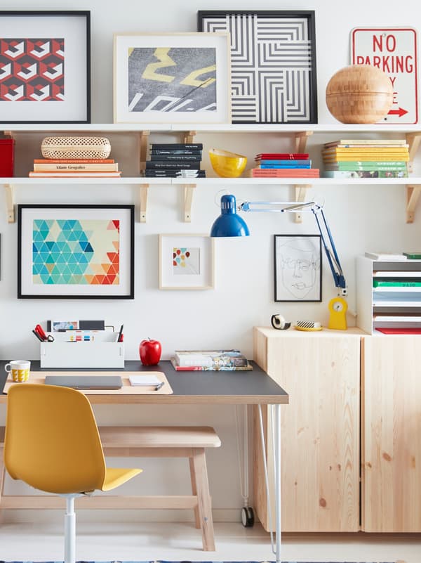 A desk on castors stands by a wall, surrounded by light-wood IVAR cabinets, wall shelves, and framed artwork.