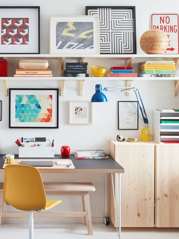 A desk on castors stands by a wall, surrounded by light-wood IVAR cabinets, wall shelves, and framed artwork.