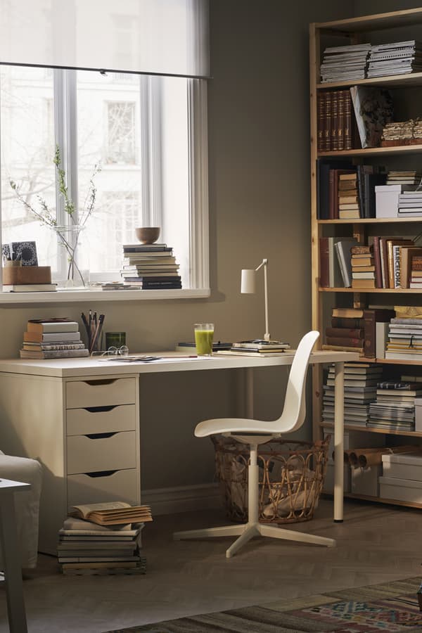 A desk space in room by a window with a white desk and work chair and bookshevles in the background.