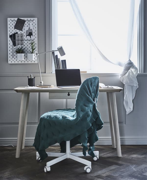 A desk with a computer and a chair with a green blanket draped over it in front of a window.