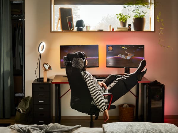 A dim room with a person reclining in a black HUVUDSPELARE gaming chair by a black HUVUDSPELARE desk with double screens.