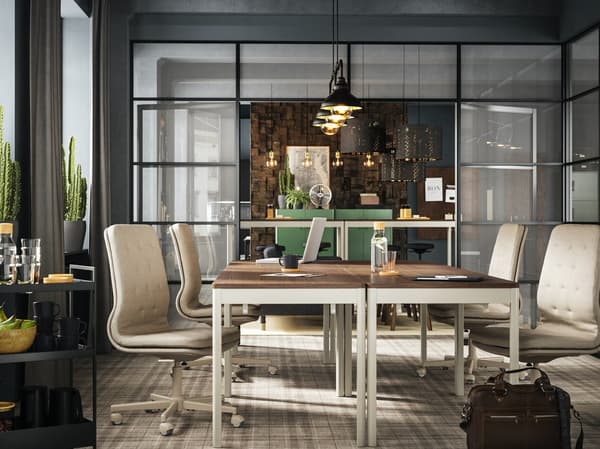 A dimly lit office meeting space featuring two wooden tables and four white swivel chairs on a carpeted flooring.