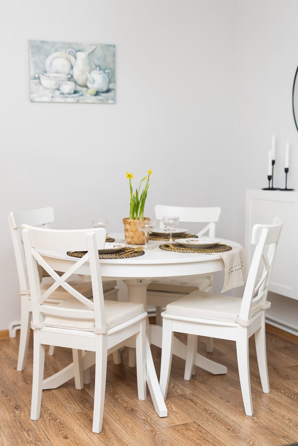 A dining area with a round table and white chairs against a light wall, with tableware and a vase of flowers placed on the table. The space looks bright and tidy.