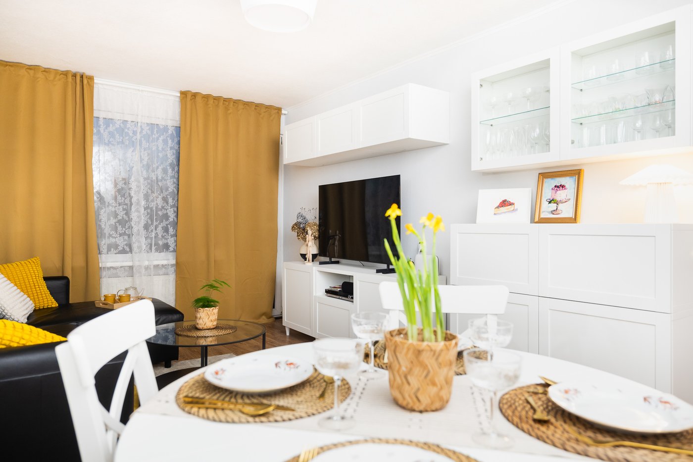 A dining area within a renovated living space featuring a round table, a white sideboard, and wall shelving. The table is set with dishes and flowers, while the living area is visible in the background.