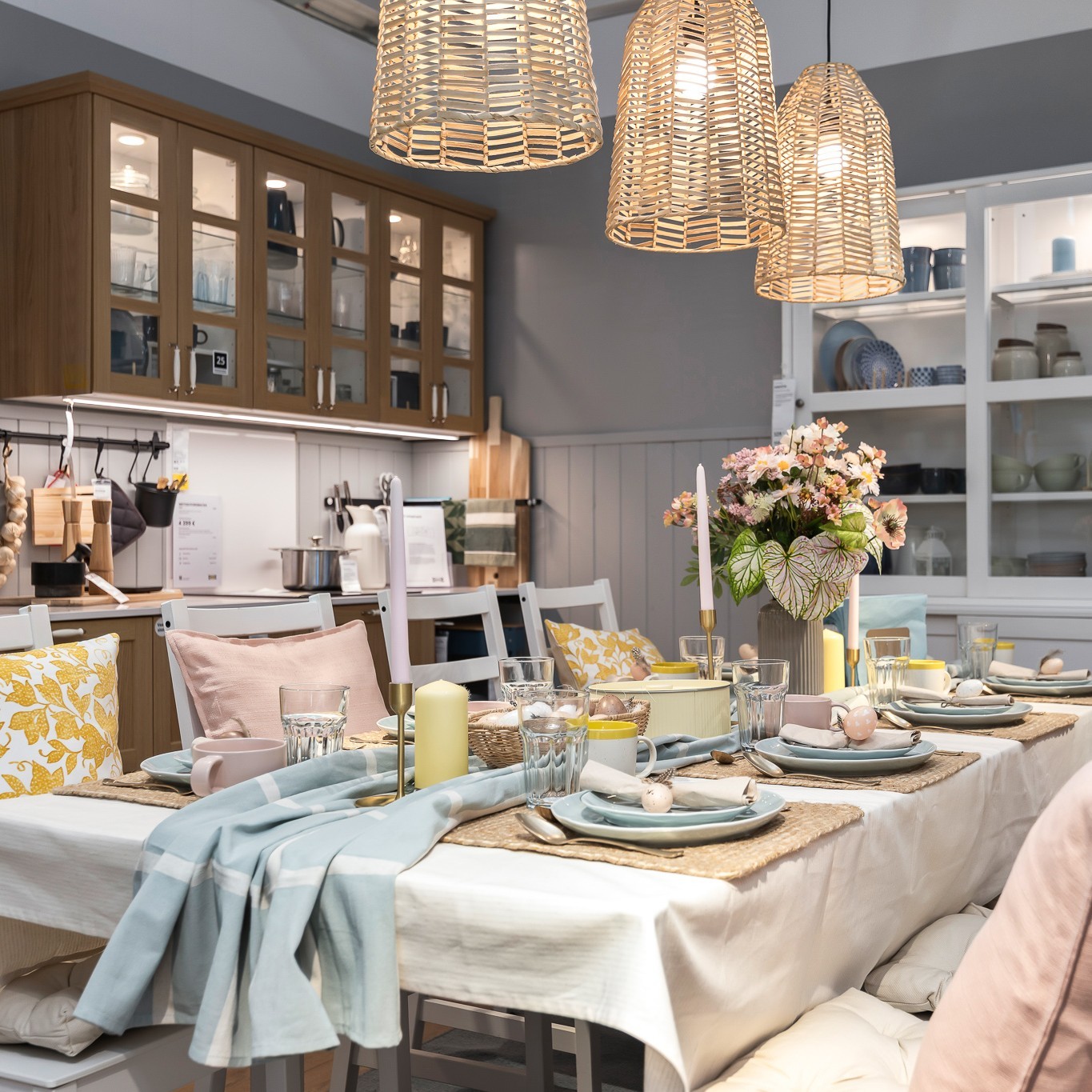 A dining room set for a festive Easter meal, featuring a table with layered textiles, pastel‑coloured plates, glassware, candles and a vase of flowers. Woven pendant lamps hang overhead, and the background shows cabinets and shelves filled with home accessories.