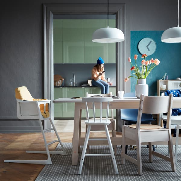 A dining room setting with a mix of chairs, leading into a kitchen, with a person sitting on the work top.
