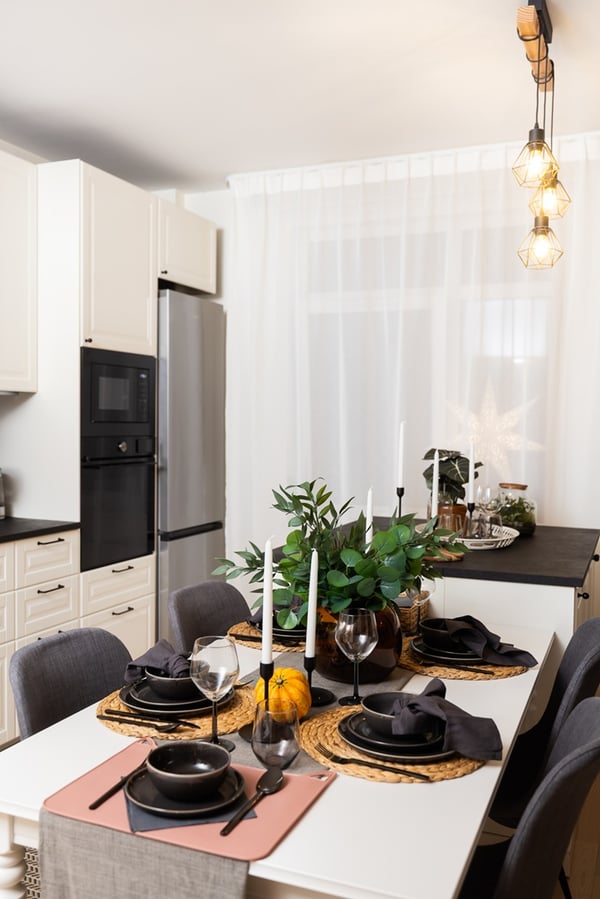A dining table in a kitchen with a dark table runner, tableware, green plants, and decorative elements, with a kitchen island in the background.