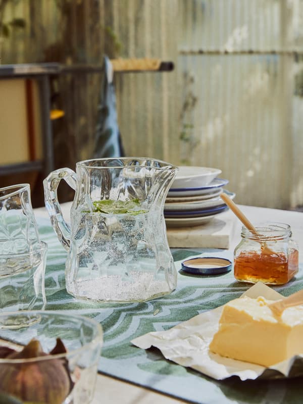 A dining table is casually set with BASTUA jug, glasses and bowl on a BASTUA bench towel used as a table runner.