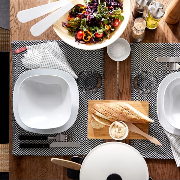A dining table, plates and bowls in white, patterned place mats, cutlery and a salad from above.