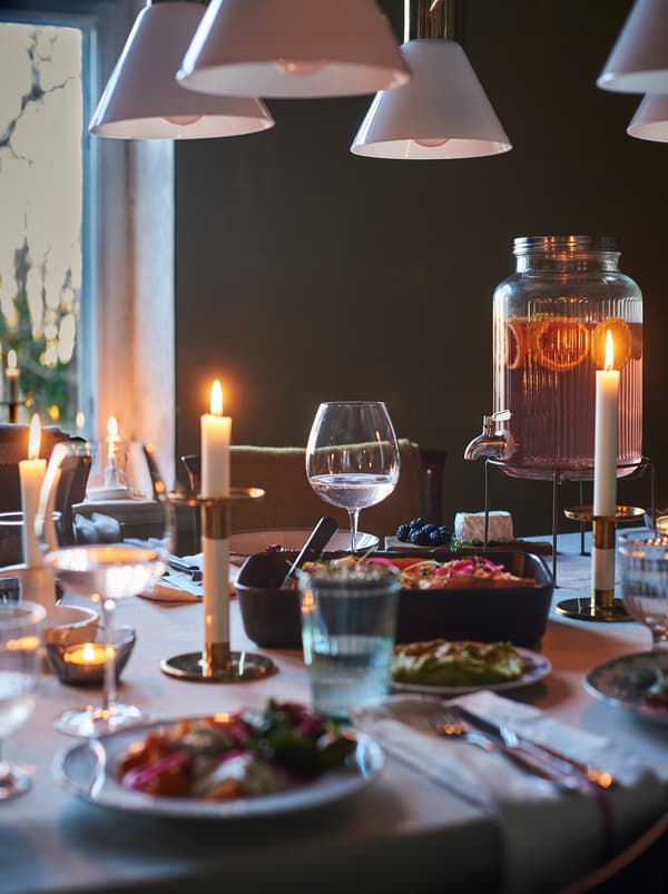 A dining table set for an informal meal with candlesticks, a beverage dispenser, pots and a roasting tin with tasty food.