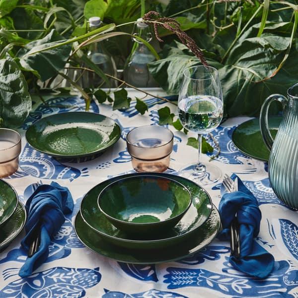 A dining table set outdoors with a green PELARKAKTUS bowl beside glassware and greenery on a floral tablecloth.