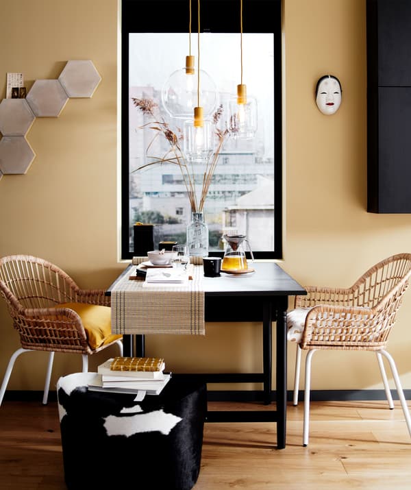 A dining table set up with two rattan chairs and a footstool in front of a window.