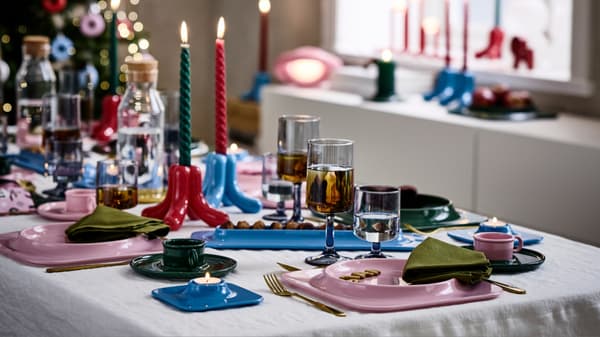 A dining table set with colorful plates, cups, napkins, candles, drinking glasses, and decorative candle holders. A lit Christmas tree is visible in the background.