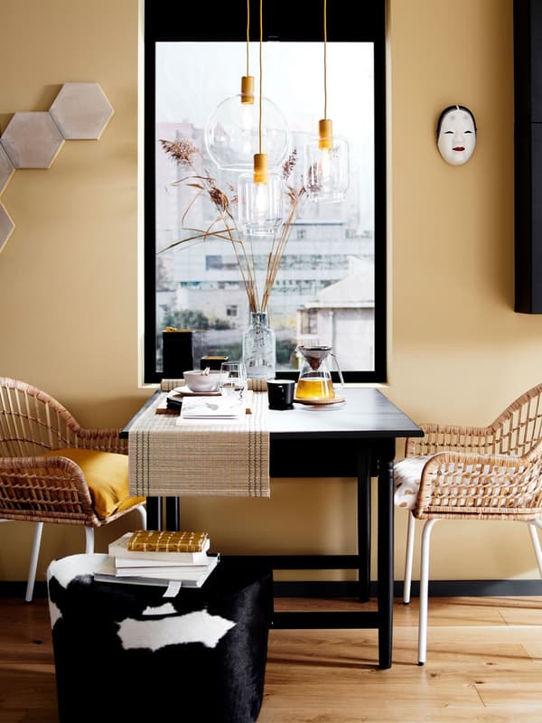 A dining table setup with two rattan chairs and a pouffe in front of a window, plus glass pendant light fittings.