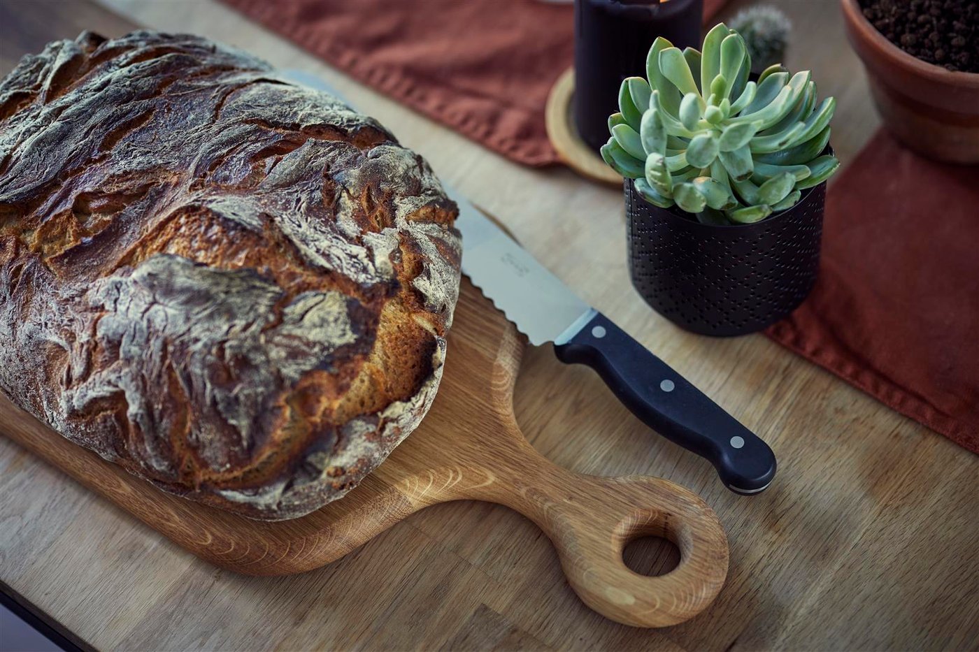 A dining table with a loaf of bread placed on a cutting board, with a bread knife next to it.