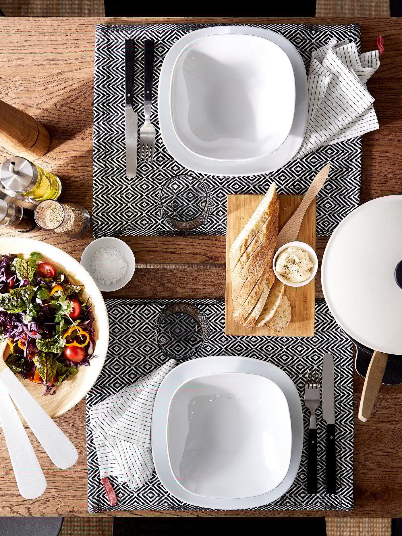 A dining table with plates and bowls in white, patterned place mats, cutlery, glasses and a salad bowl.