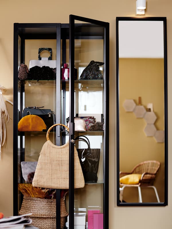 A display cabinet containing a variety of objects including handbags stands next to a mirror which is mounted on a wall.