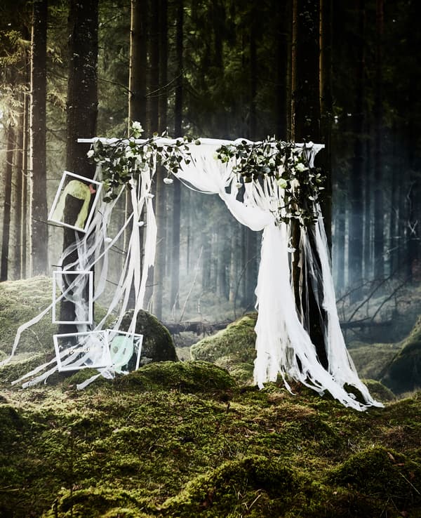 A DIY photobooth backdrop made from strips of lacy curtains and fake flowers secured to a pole that’s hanging up in a forest wedding