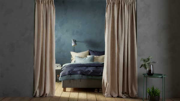 A doorway with open beige AINA curtains hanging in it. Beyond the doorway is a bed with white and blue bed linen.