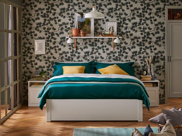 A double bed with green bedding and yellow cushions. A shelf is behind it on the wall and a pendant lamp hangs above.