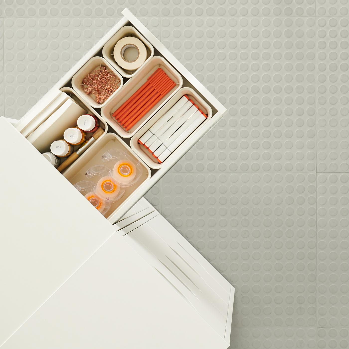 A drawer sticking out from a white unit holding different organizers with tape, pens and paper clips all with orange accents.