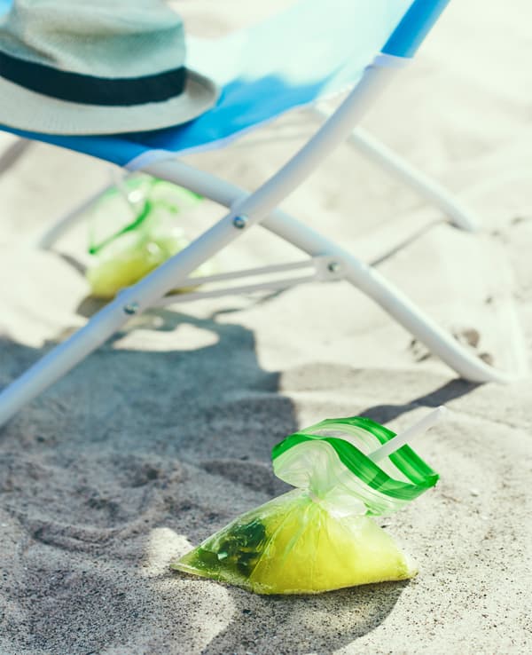A drink served in a plastic bag sits in the sand next to a beach chair.