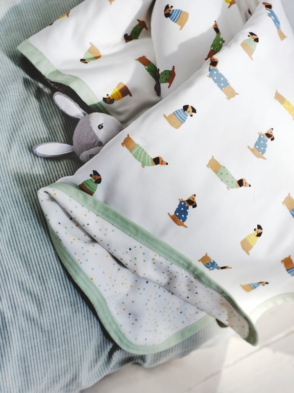 A DRÖMSLOTT blanket in patterns of puppies covers a GULLIGAST rabbit soft toy on a bed covered with BERGPALM duvet.