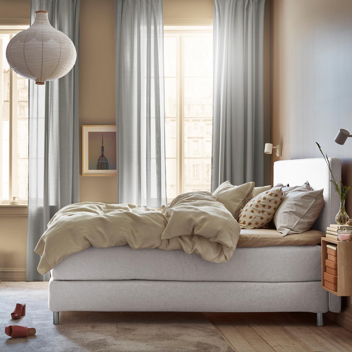 A DUNVIK divan bed stands in a bright bedroom with two long windows and a pair of shoes lying on the rug.