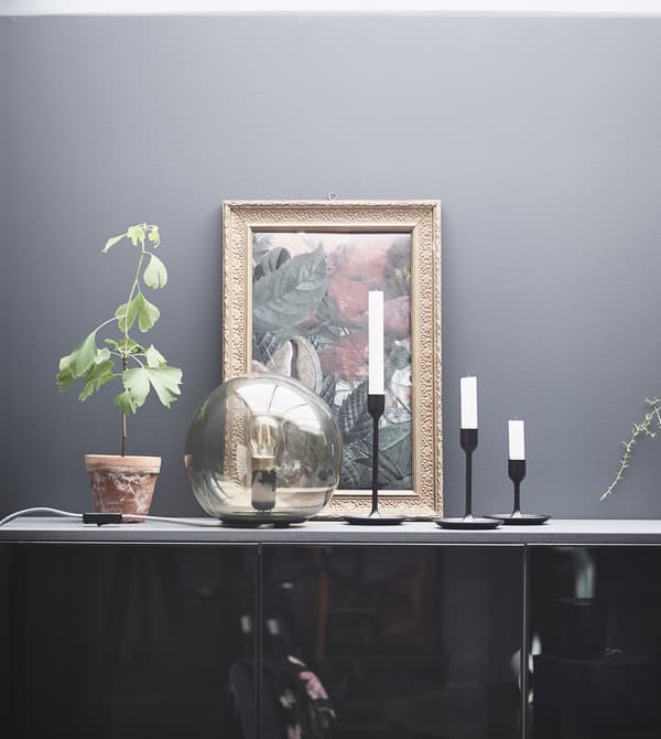 A FADO table lamp adds ambience to a bedroom display with candles, a painting, and plant on a cabinet.