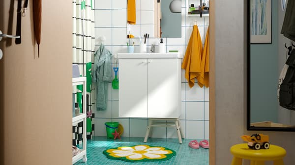A family bathroom with white ENHET furniture and a green floor with a KÄRRKNIPPROT rug, and wall hooks at different heights.