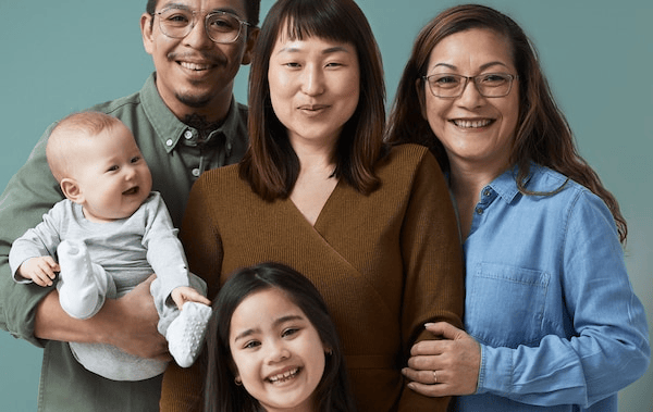 A family of five, all smiles from couple with a baby, a young daughter and a grandmother standing close together.