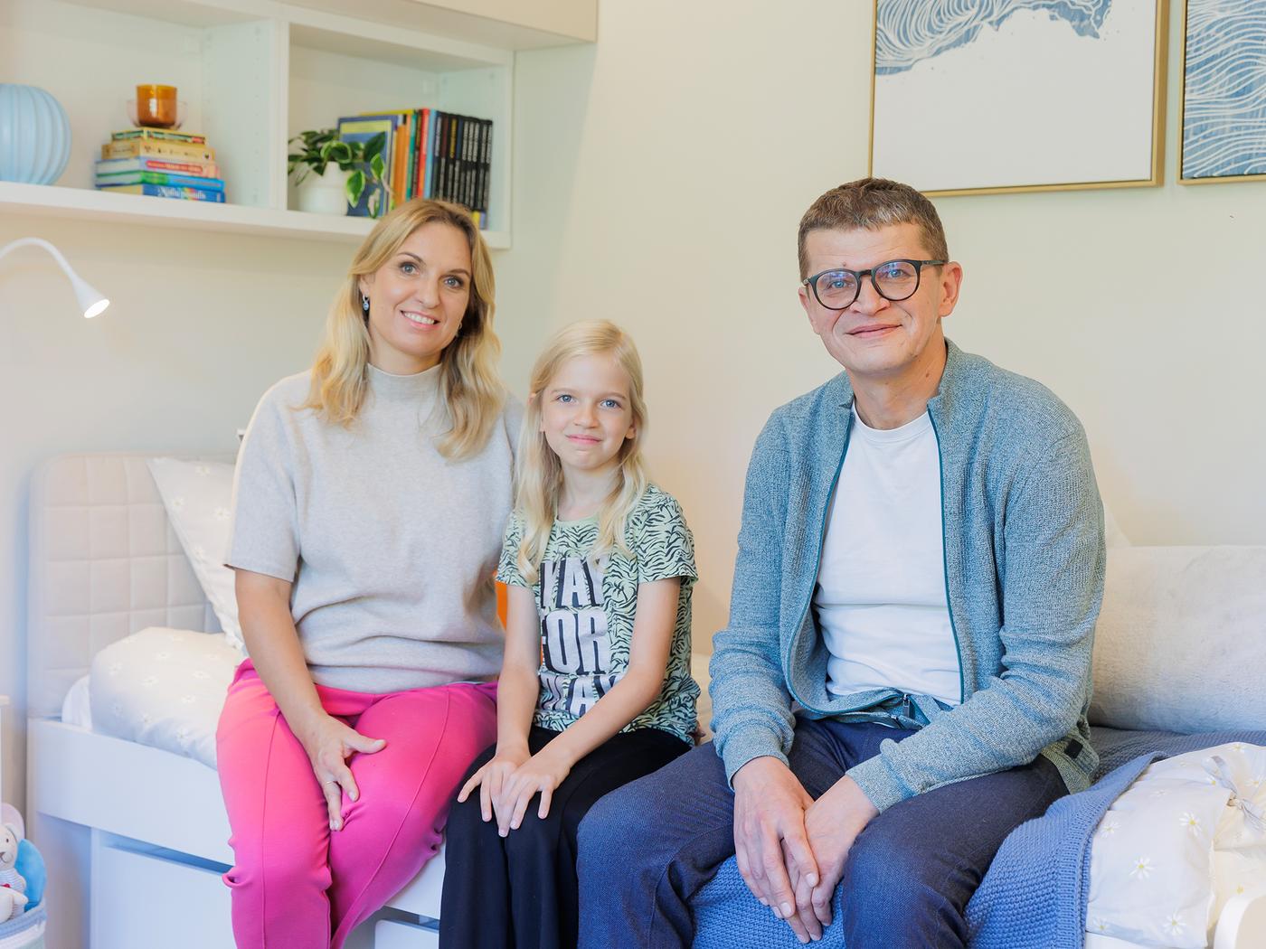 A family of three sits together on a bed in a cosy, well-lit room. The background shows a neatly arranged bookshelf and framed artwork on the wall.