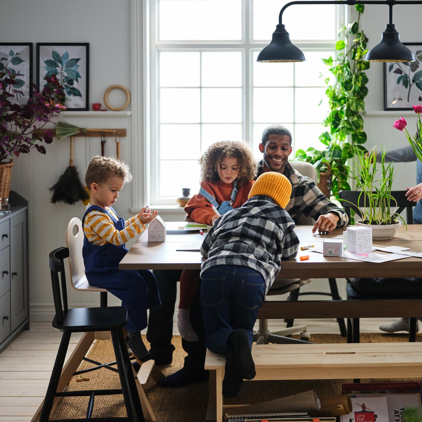 A family of three small children and a father seated at a wooden dining table with a windowed backdrop and potted plants.