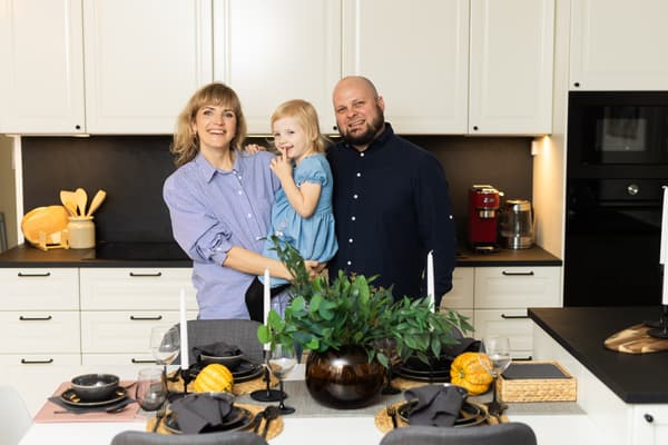 A family of three stands in a modern kitchen: a woman holding a young child with a man beside her. In the foreground is a dining table with black tableware, pumpkins, and green plants.