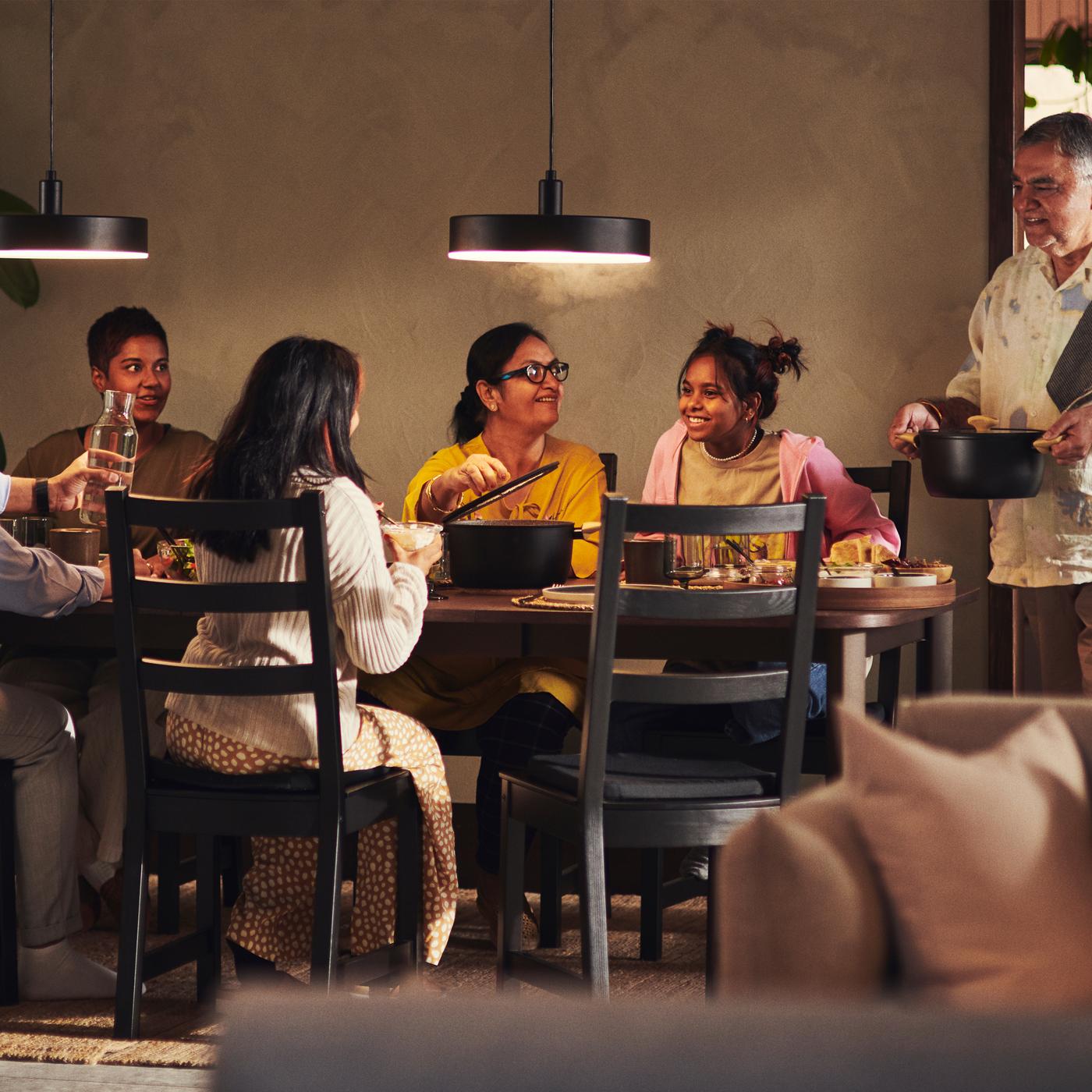 A family sits together at a STRANDTORP dining table. A person stands ready to serve food from a HUSKNUT pot with lid.