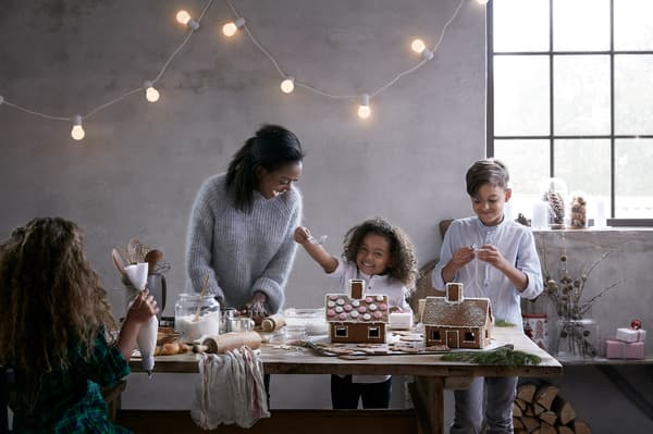 A family stands around a large harvest table that is filled with supplies to make and decorate gingerbread cookies, including gingerbread houses.