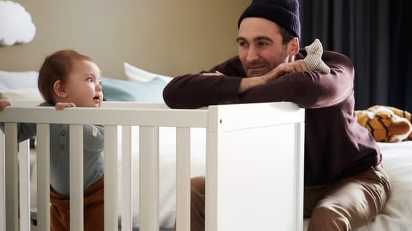 A father plays with his baby and a small soft toy as the baby stands in a white SUNDVIK cot holding on to the side.