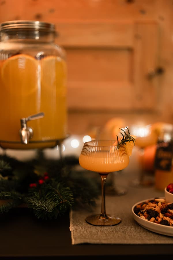 A festive cocktail scene with a glass of orange beverage garnished with rosemary, beside a jar of punch. Warm lighting and holiday decor create a cozy atmosphere.