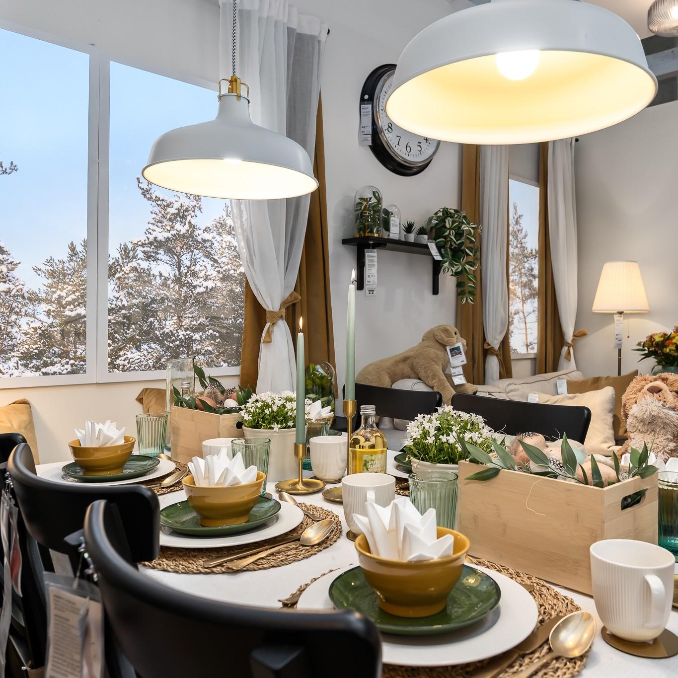 A festive Easter table set with layered green and yellow plates and bowls, folded napkins, candles and small natural arrangements, with large windows showing a snowy landscape in the background.