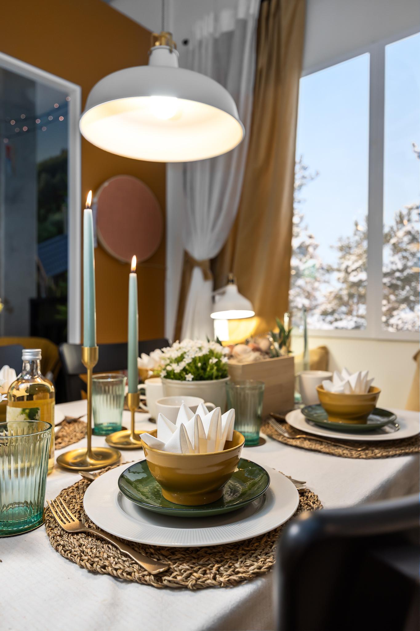 A festive Easter table set with layered plates and bowls in earthy yellow and green tones, folded napkins arranged in bowls, tall candles, and glassware.