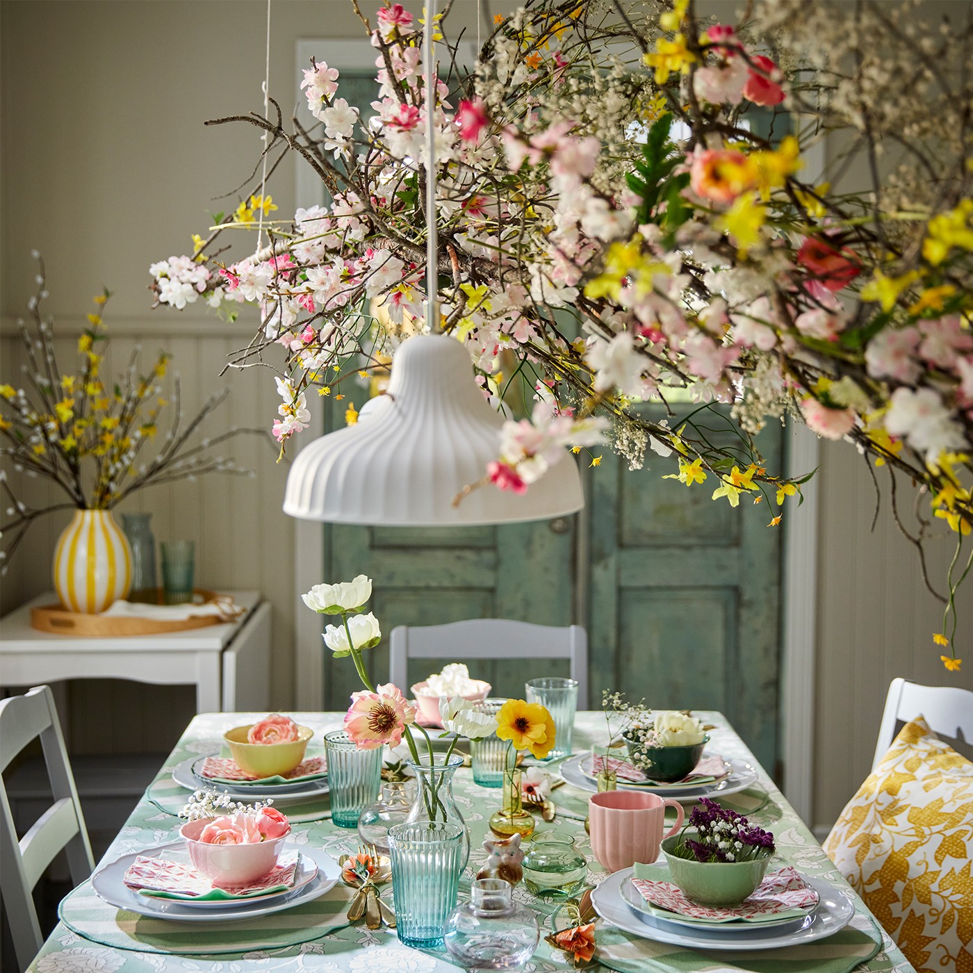 A festive spring table setting with a stack of bowls in yellow, dark green, pink and light green, placed on top of side plates and plates, and decorated with artificial spray magnolias.