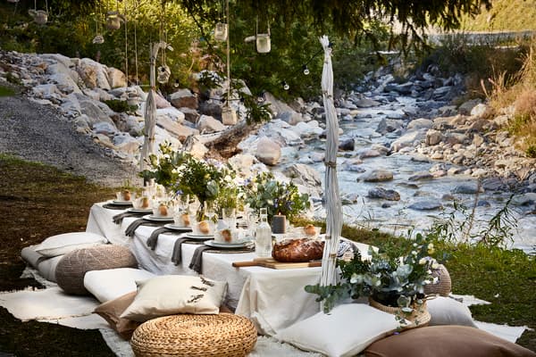 A festively decorated table with a white table cloth, flowers in vases and cushions and poufs next to it.