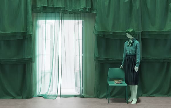 A figure dressed in green standing next to a green chair with a backdrop of green layered curtains.