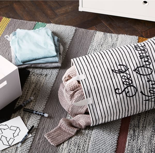 A filled striped KLUNKA laundry bag and stack of clothing on a grey carpet.