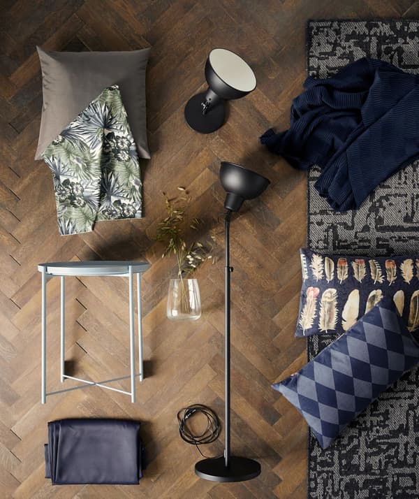 A flatlay featuring a black SKURUP lamp, various cushions, a modern grey and black carpet, a light blue GLADOM tray table, and assorted bric-a-brac, all arranged on a brown wood herringbone floor