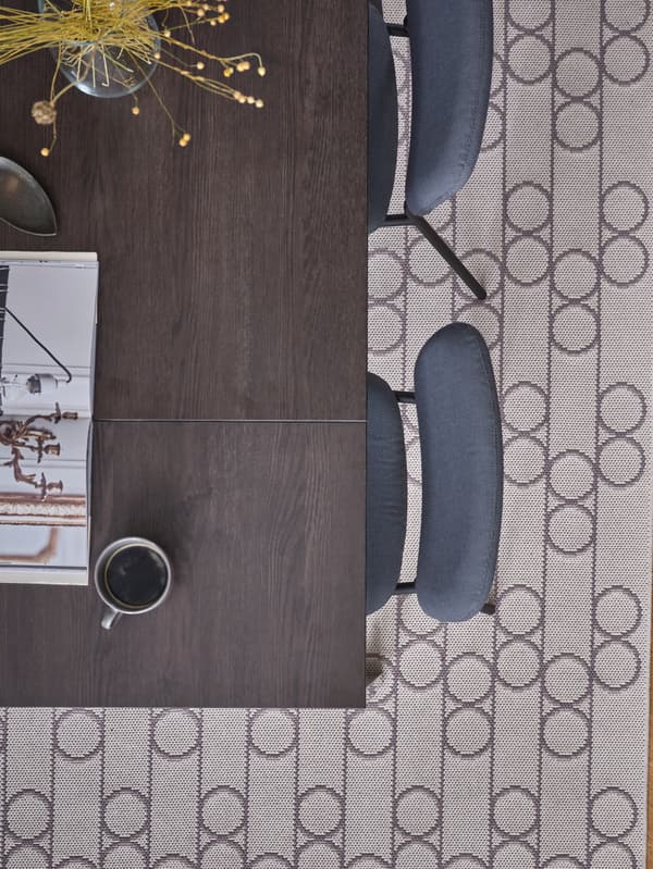 A flatwoven rug with a geometric pattern with circles lies beneath a black/dark brown dining table and grey chairs.
