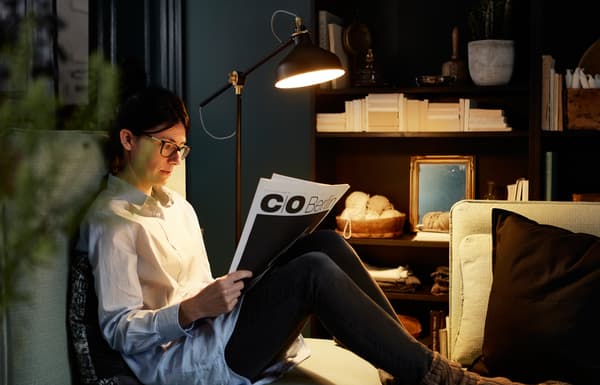 A floor lamp lights a newspaper for a woman reading on the sofa.