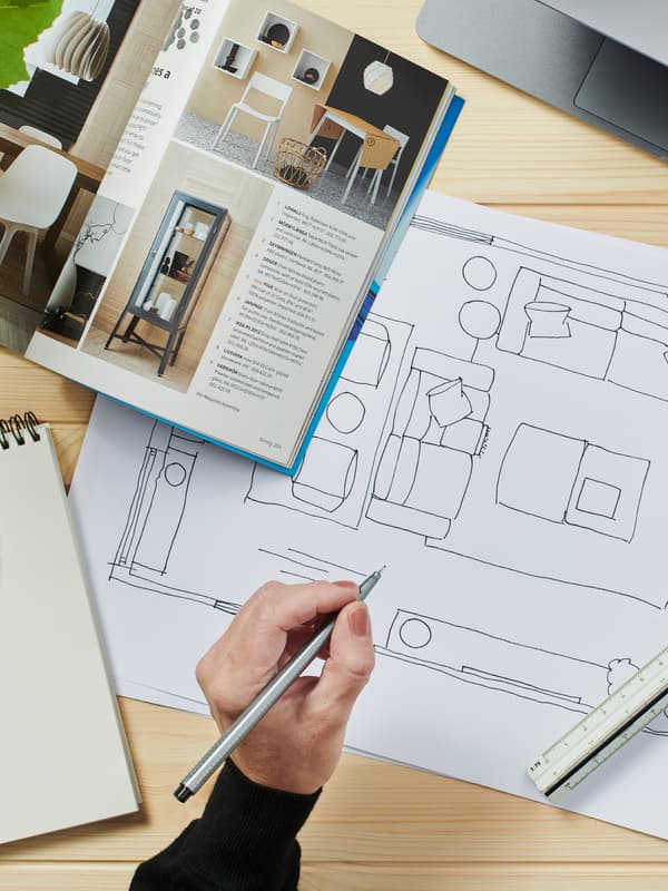 A floorplan and IKEA catalog, with a hand holding a pencil above a work desk.