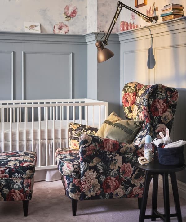 A floral armchair and footstool next to a baby’s cot, with clamp light above.